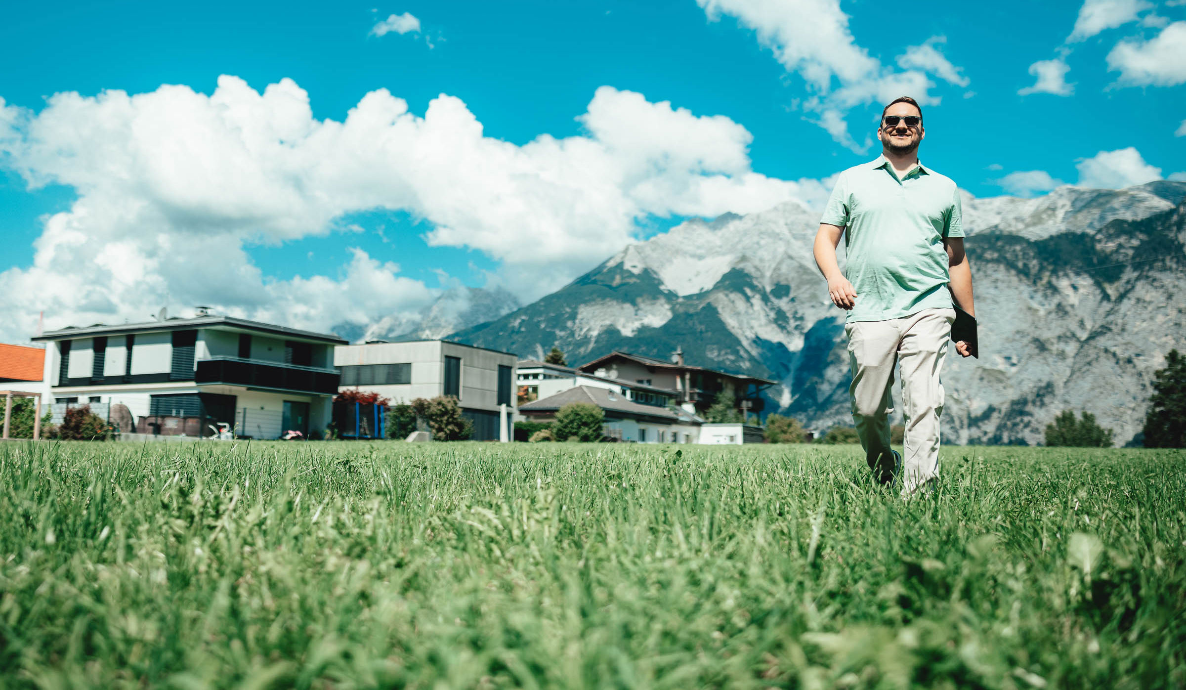 Immobilienberater Fabian geht mit Tablet über ein Grundstück vor alpiner Bergkulisse