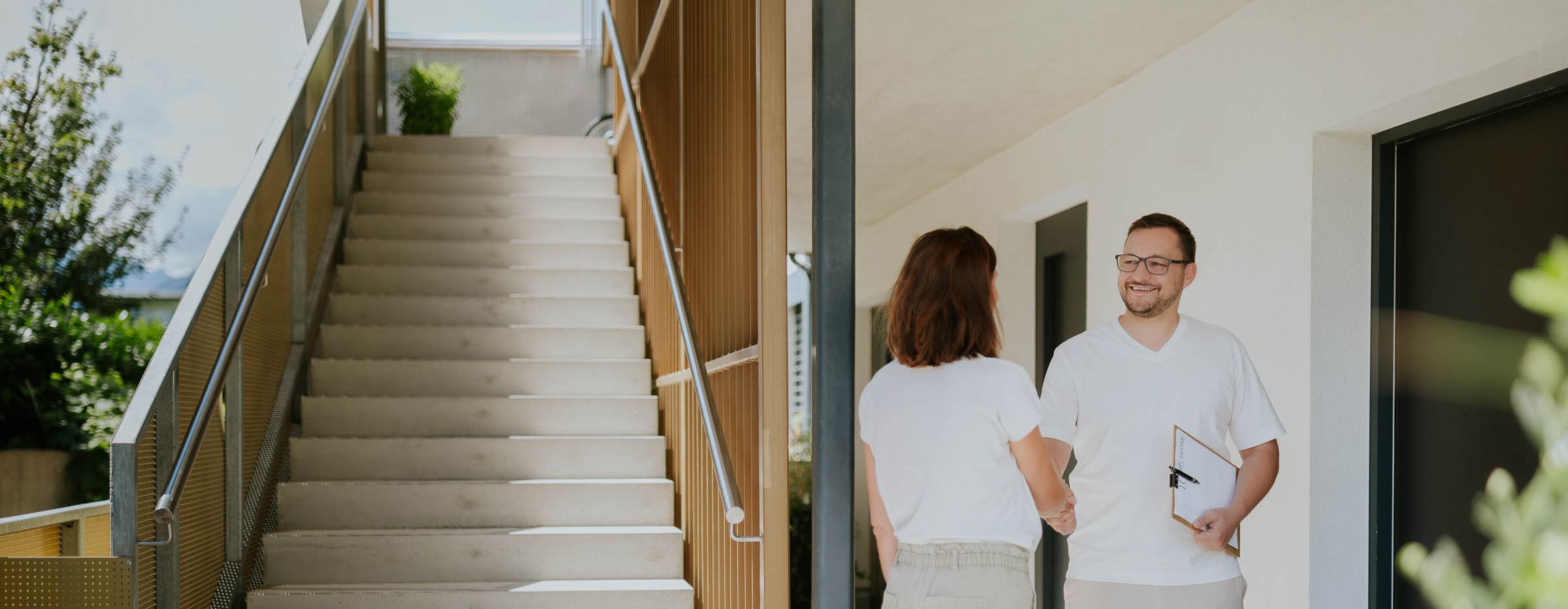 Immobilienberater Fabian begrüßt eine Kundin mit Händedruck vor einem modernen Gebäude